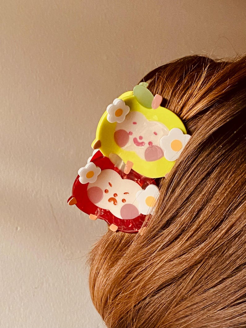 May include: Two cartoon apple-shaped hair clips. One is yellow with a green leaf and white flower accents, the other is red with white flower accents. Both have smiling faces and are set against brown hair.