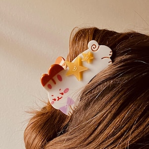 May include: A white and orange cat-shaped hair clip with yellow star accents. The clip features a cartoon cat design with a pink bow tie and a red and orange headpiece. The hair accessory is set against brown hair.