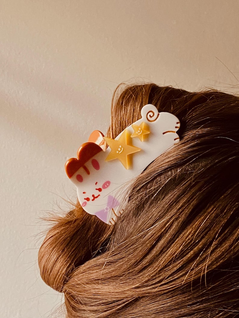 May include: A white and orange cat-shaped hair clip with yellow star accents. The clip features a cartoon cat design with a pink bow tie and a red and orange headpiece. The hair accessory is set against brown hair.