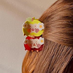 May include: Two apple-shaped hair clips, one yellow and one red, are clipped into brown hair. Each clip features a smiling face and white flower accents. The yellow clip is on top, and the red clip is below.