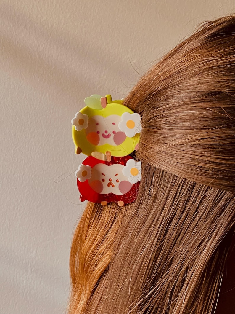 May include: Two apple-shaped hair clips, one yellow and one red, are clipped into brown hair. Each clip features a smiling face and white flower accents. The yellow clip is on top, and the red clip is below.