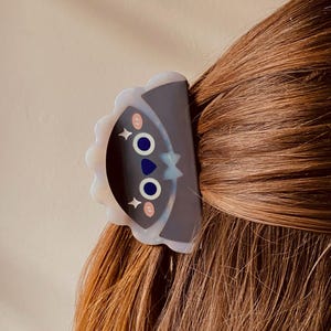 May include: A decorative hair clip featuring a cartoon face design with blue eyes, pink cheeks, and a star. The clip is dark gray with a scalloped edge and is attached to brown hair.