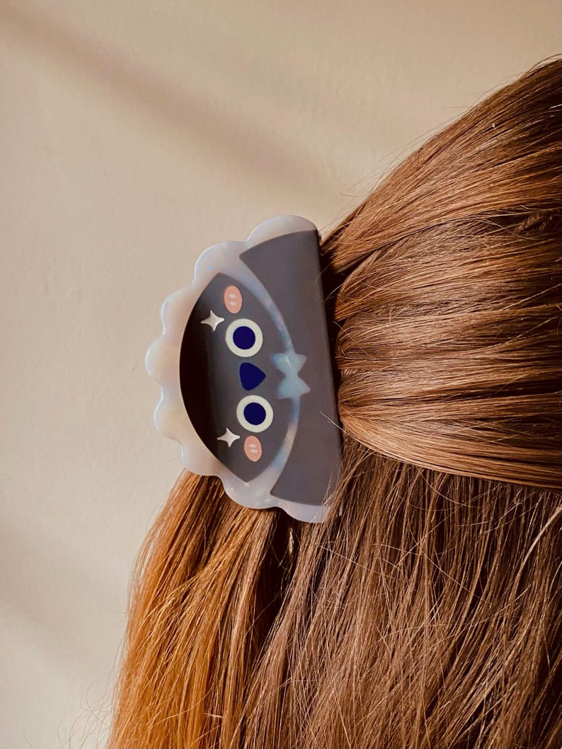 May include: A decorative hair clip featuring a cartoon face design with blue eyes, pink cheeks, and a star. The clip is dark gray with a scalloped edge and is attached to brown hair.