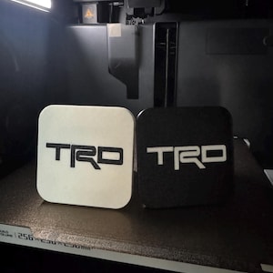 Puede incluir: Dos posavasos cuadrados con esquinas redondeadas, uno blanco con texto negro y otro negro con texto blanco, ambos con las letras TRD en una fuente estilizada. Los posavasos están colocados dentro de una impresora 3D con base visible.