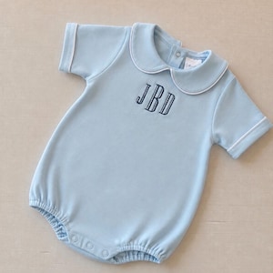 May include: A light blue short-sleeve romper featuring a rounded Peter Pan collar with white piping trim. The garment includes a dark blue embroidered monogram with the letters JBD on the chest and snap closures at the bottom and back neck.