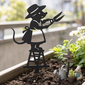 May include: A black metal garden stake silhouette of a mouse wearing a hat and apron, standing on a small stool while holding pruning shears. The design features a pointed base for insertion into soil, depicting a gardening scene in a flat, cutout style.
