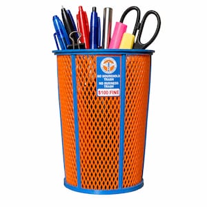Op de afbeelding: Een oranje pennenbak van gaas met een blauw frame, gevuld met pennen, een schaar en een paperclip. Een rechthoekig bordje aan de voorzijde vermeldt: NEW YORK CITY DEPARTMENT OF SANITATION, NO HOUSEHOLD TRASH, NO BUSINESS TRASH, $100 FINE.