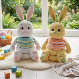 May include: Two crochet bunny stacking toys. One is white with blue and yellow rings, the other is yellow with orange rings. Both have long ears, black eyes, and pink noses. They sit on a white rug with wooden blocks and a basket of yarn in the background.