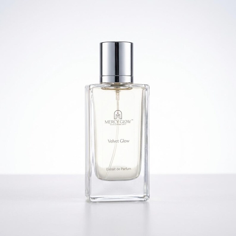 May include: A rectangular glass perfume bottle with a silver cap. The bottle contains a clear liquid and has the text "MERCY GLOW" and "Velvet Glow" printed on the front. The words "Extrait de Parfum" are also printed on the bottle.