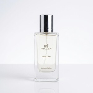 May include: A rectangular perfume bottle with a silver cap. The bottle is clear, revealing a light-colored liquid inside. The label reads "MERCY GLOW" above "Velvet Glow" and "Extrait de Parfum."
