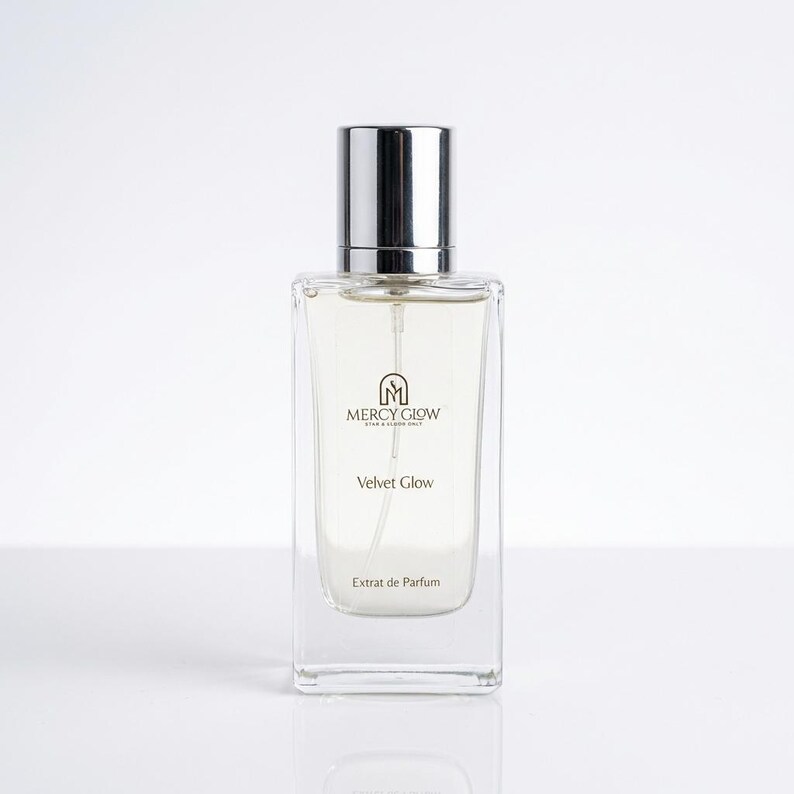 May include: A rectangular perfume bottle with a silver cap. The bottle is clear, revealing a light-colored liquid inside. The label reads "MERCY GLOW" above "Velvet Glow" and "Extrait de Parfum."
