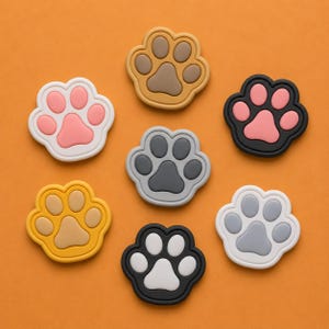 May include: An assortment of colorful paw-shaped accessories in various colors, including pink, brown, gray, yellow, and black, with white or black outlines. The accessories are arranged on an orange background.