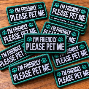 May include: A collection of black rectangular patches with the text "I'M FRIENDLY PLEASE PET ME" in white. The patches have a teal border and teal paw prints. The patches are stacked and scattered on a wooden surface.