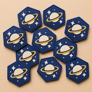 May include: A collection of navy blue, hexagon-shaped coasters. Each coaster features a cream-colored planet with a yellow ring, surrounded by white stars. The coasters are designed with a space or galaxy theme.