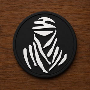May include: A circular black patch featuring a white graphic design of a figure wearing a head wrap and draped clothing. The design is centered on the matte black surface, creating a high-contrast visual of a stylized person in traditional attire.