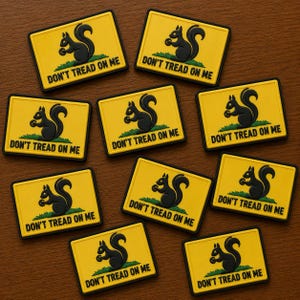 May include: A collection of yellow rectangular patches with a black border. Each patch features a black squirrel on green grass, with the text "DON'T TREAD ON ME" in black. The patches are arranged on a brown wooden surface.
