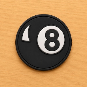 May include: A black and white eight ball coaster. The coaster is round with a black background and a white number eight in the center. A white triangle shape is on the left side of the eight ball.