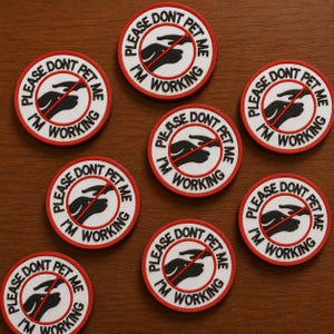 May include: A collection of round, red-rimmed stickers with the text "PLEASE DON'T PET ME I'M WORKING" in black and white. The design includes a hand with a red line through it, indicating "no petting."