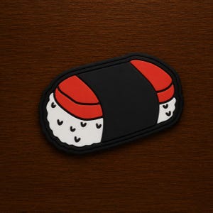 May include: A sushi-shaped patch with a black border. The sushi has a black center, red topping, and white rice with black accents. The patch is on a dark brown wooden surface.