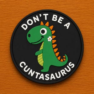May include: A round, black coaster with the text "DON'T BE A CUNTASAURUS" in white. The coaster features a cartoon green dinosaur with orange spikes and a white flower. The background is a wood grain pattern.