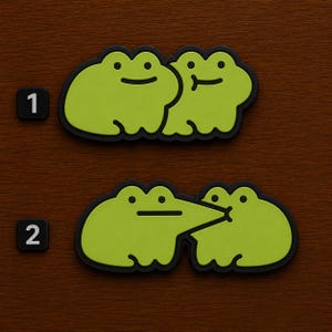 May include: Two sets of green frog-shaped stickers with black outlines. One set shows two frogs side-by-side, and the other shows two frogs kissing. The number "1" and "2" are in black squares.