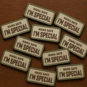 May include: A collection of rectangular patches with the text "MAMA SAYS I'M SPECIAL" in a dark brown font. The patches have a light beige background and a dark green border. The patches are scattered on a brown wooden surface.