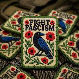 May include: Rectangular patch with the text "FIGHT FASCISM" above a black crow with blue wings, surrounded by red flowers and green leaves. The patch has a green border and is set against a patterned background.