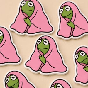 May include: A collection of cartoon frog-shaped stickers. Each sticker features a green frog with large eyes, wearing a pink blanket. The stickers have a white border and are arranged on a light beige background.