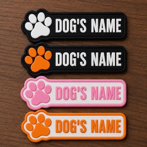May include: Four personalized dog name tags in black, orange, pink, and white. Each tag features a paw print design and the text "DOG'S NAME" in bold, white letters. The tags are displayed on a wooden surface.