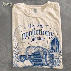 Pode incluir: Uma t-shirt cor de marfim com uma impressão gráfica azul com o texto 'it's too nonfictiony outside'. O design inclui uma lua crescente, um gato a dormir sobre uma pilha de livros com as etiquetas 'Fantasy', 'Magic', 'Good Books' e 'Happier Days'.