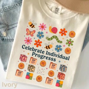 Könnte beinhalten: Ein elfenbeinfarbenes T-Shirt mit einem bunten Grafikdruck aus Insekten, Blumen und pädagogischen Symbolen wie Alphabet-Blöcken, Puzzleteilen und mit IEP beschrifteten Ordnern. Der Text Celebrate Individual Progresss ist in Dunkelblau gedruckt.