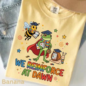 May include: A pale yellow t-shirt featuring a graphic of a bee, a frog in a red cape, and a snail, all wearing graduation caps and rainbow scarves. They hold clipboards with checklists. The text reads WE REINFORCE AT DAWN in rainbow colors.
