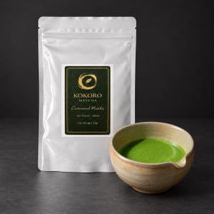 Könnte beinhalten: Ein weißer, wiederverschließbarer Beutel Kokoro Matcha Ceremonial Matcha aus Uji, Kyoto, Japan, mit einem Nettogewicht von 50g, neben einer hellen Keramikschale, die mit leuchtend grünem Matcha-Tee gefüllt ist.