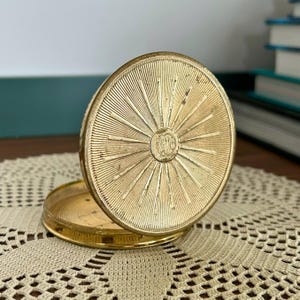 May include: A round gold-tone metal compact case with a hinged lid. The lid features a sunburst pattern of radiating lines emanating from a central circular medallion. The base is also round and gold-toned, matching the lid's metallic finish.