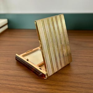 May include: A square compact case featuring a gold-toned metal lid with a vertical pinstripe texture. The base of the compact is constructed from a dark brown tortoiseshell-patterned material, and the interior contains a rectangular mirror.