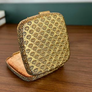 May include: Open, antique gold-toned compact with a detailed, repeating fan-shaped pattern. The compact is square with a hinged lid and a peach-colored powder puff inside. The compact is open, revealing the powder and mirror.
