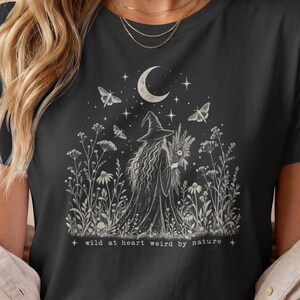 以下が含まれることがあります： ダークグレーのTシャツ。マントと帽子を身につけた魔女が、三日月と星の下、野花が咲く野原に立っている白いグラフィックプリントが施されており、イラストの下には「wild at heart weird by nature」というテキストが印刷されています。