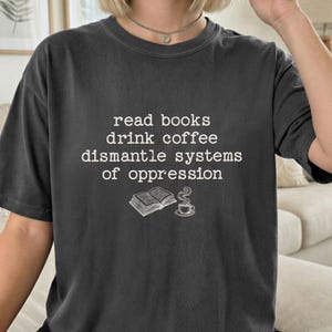 Puede incluir: Camiseta gris oscuro con el texto blanco "read books drink coffee dismantle systems of oppression". Una ilustración de un libro y una taza de café debajo. Camiseta de estilo casual y unisex.