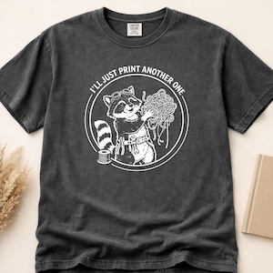 以下が含まれることがあります： ゴーグルをかけたアライグマがフィラメントの塊を持っている白いグラフィックがプリントされた、ペッパーカラーのTシャツ。「I'll just print another one」の文字がアライグマの周りに円状にプリントされています。