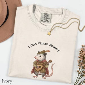 Può includere: T-shirt color avorio con la scritta "I Cast Vicious Mockery" e un disegno di un opossum che suona un liuto. L'opossum indossa un cappello e un mantello. Sono visibili anche una collana a stella dorata e un cappello marrone.