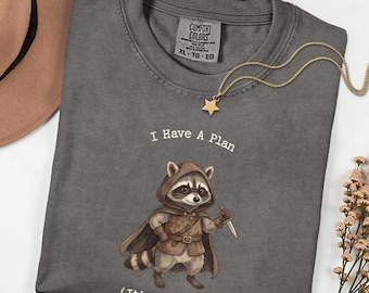May include: Pepper-colored Comfort Colors t-shirt with the text "I Have A Plan (It's A Bad One)" and a cartoon raccoon wearing a cloak and holding a knife. A gold star pendant necklace is draped across the shirt.
