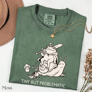 May include: Moss green Comfort Colors t-shirt with a whimsical illustration of a frog riding a snail, holding a sword. The text "TINY BUT PROBLEMATIC" is printed below the graphic. A gold star pendant necklace is draped on the shirt.