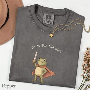 May include: A dark gray t-shirt with the phrase "Do it for the plot" and a cartoon frog holding a sword. The frog wears a cape. A gold star necklace and a brown hat are also visible.