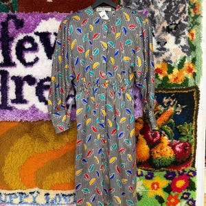 May include: A vintage dress with a black and white striped pattern, adorned with colorful leaf-like shapes in red, yellow, blue, and green. The dress features long sleeves, a button-down front, and a cinched waist. The dress is hanging on a black hanger.