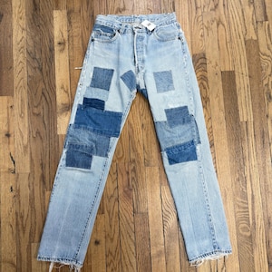 Può includere: Jeans in denim azzurro chiaro con taglio a gamba dritta e chiusura a bottoni. I pantaloni sono decorati con molteplici toppe rettangolari in varie tonalità di blu scuro e medio, e gli orli delle gambe presentano una finitura sfrangiata.
