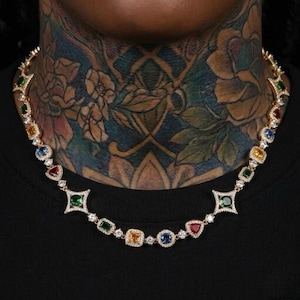 May include: A gold-toned necklace featuring a series of colorful gemstones in various shapes, including emerald green, sapphire blue, ruby red, and citrine yellow. The necklace is set with small, clear accent stones and is worn against a black background.