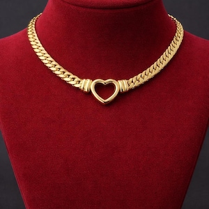 May include: A gold-colored necklace with a heart-shaped pendant. The necklace features a thick, flat chain design. The heart pendant is open in the center. The necklace is displayed on a red velvet bust.