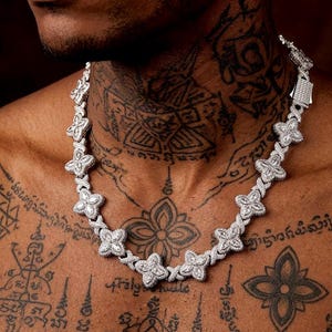 May include: A silver-colored necklace featuring a floral design. The necklace is composed of multiple flower-shaped links, each adorned with small, sparkling stones. The necklace is worn around the neck and chest.