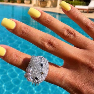 May include: A silver-toned ring shaped like a cat face, fully encrusted with clear pavé stones. The design features two dark oval eyes, a small yellow nose, and three whiskers on each side, all set against a bright, sparkling metallic surface.