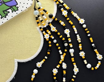 May include: A phone charm strap featuring a repeating pattern of black, yellow, and white beads interspersed with small white daisy flower beads. The strap is attached to a white phone case decorated with a green cartoon alligator illustration.